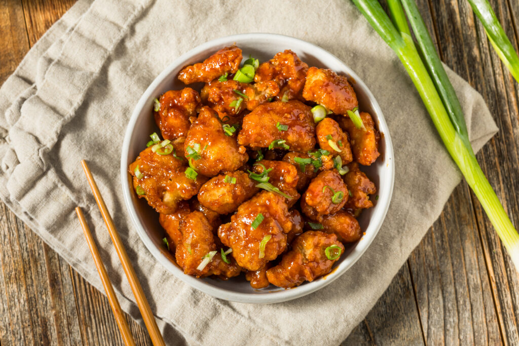 Crispy glazed chicken with green onions and sweet chili sauce served in a white bowl with chopsticks